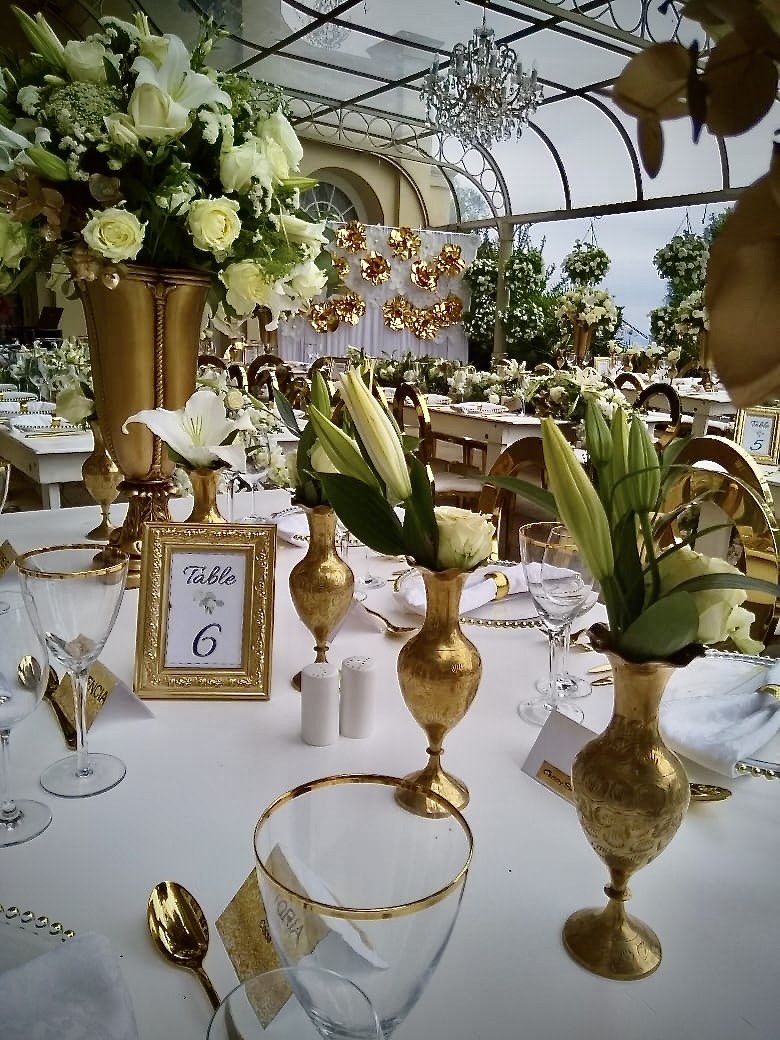 Wedding tablescape styled by Memories in Motion with gold vases, white lilies and roses, gold-rimmed glassware and framed table numbers under a conservatory chandelier